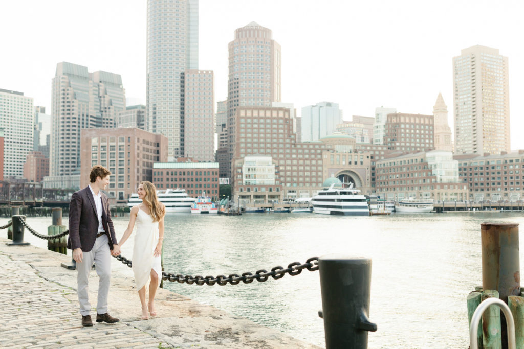 a couple doing their engagement photos in boston's seaport area. he's wearing a jacket and she's in a white dress.