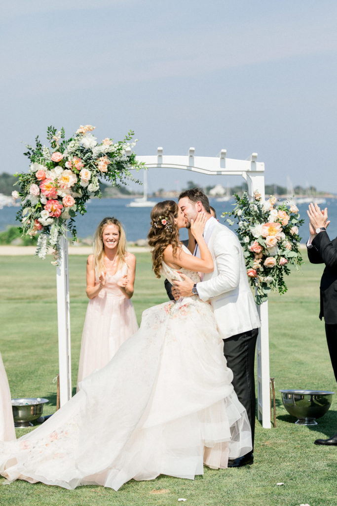 First kiss at Wentworth by the Sea Country Club Rye NH wedding, florals by Jardiniere, Lazaro floral bride's gown, Indochino groom's white tux