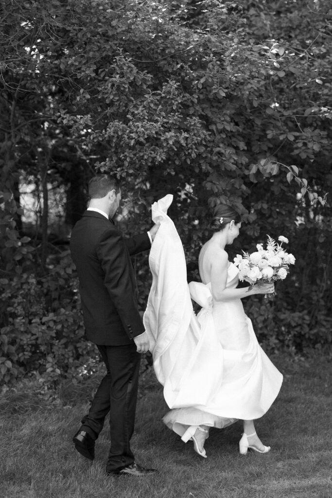 Black and white candid portrait of bride and groom walking together and the groom helping with her dress
